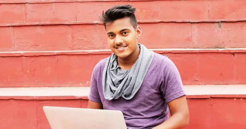Smiling young man in a purple t-shirt and grey scarf sitting on red steps with a laptop