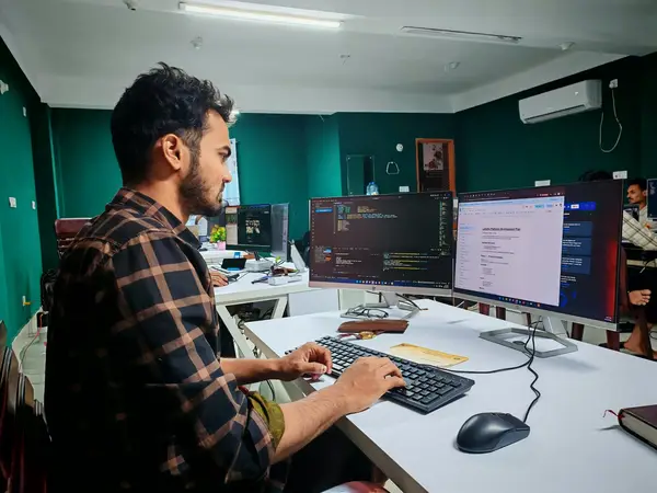 Man in a checked shirt typing at a desk with two monitors showing code and a document in an office
