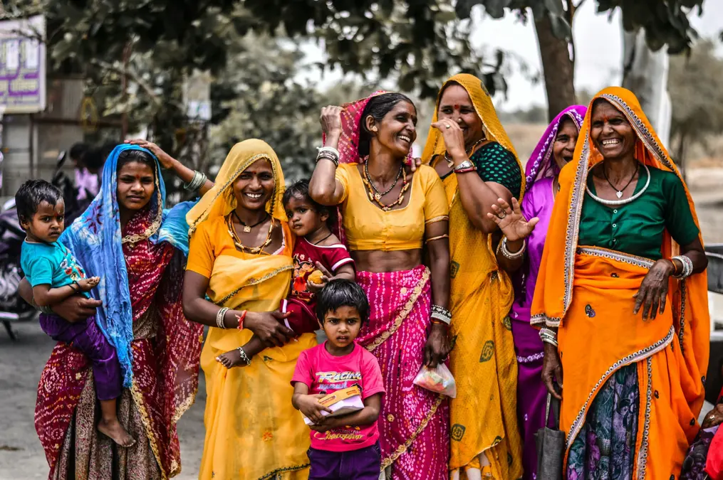 Group of laughing women in bright saris with young children standing by a village road