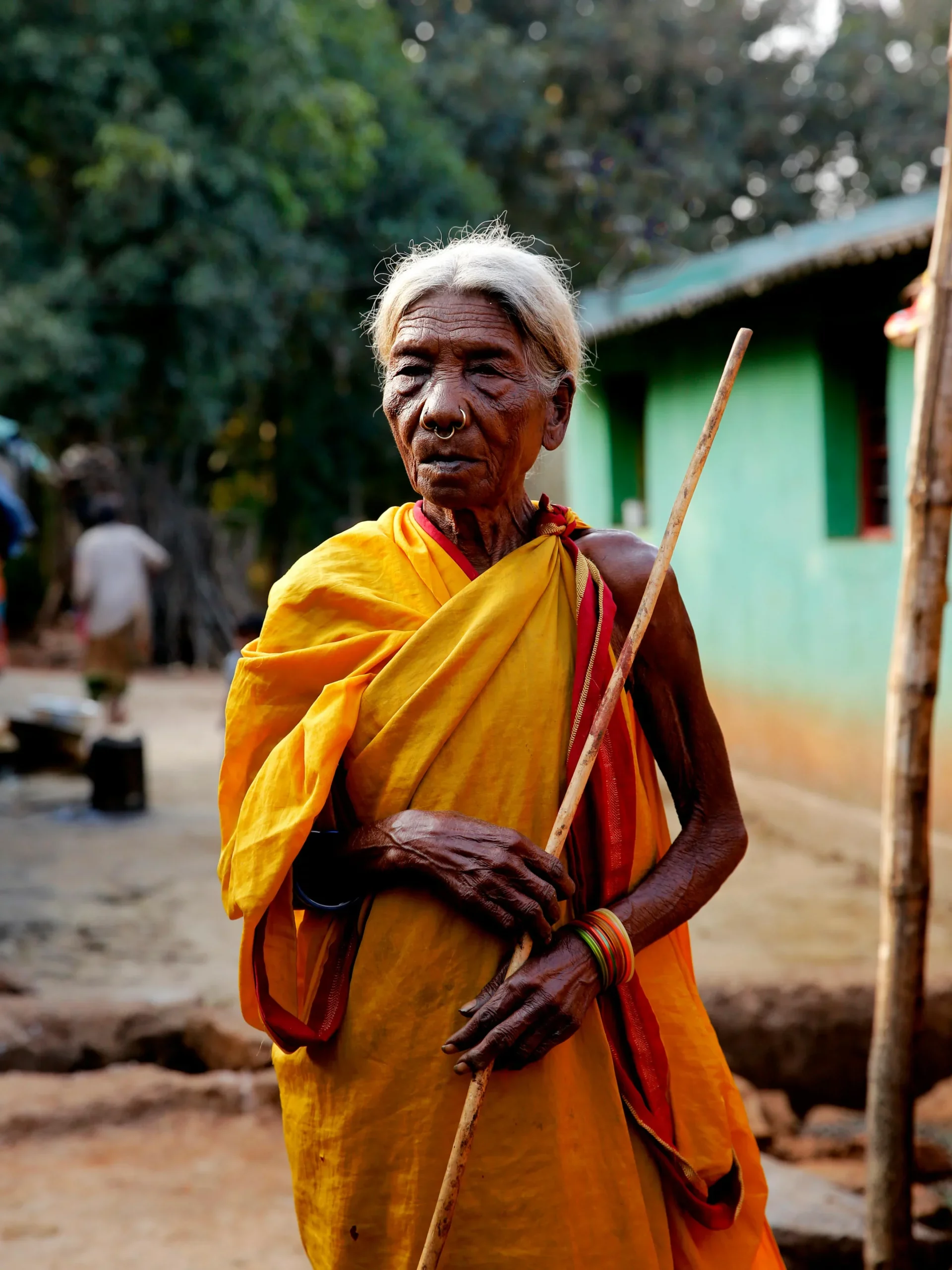 Elderly woman in a yellow sari holding a wooden stick in a village with a green house behind her