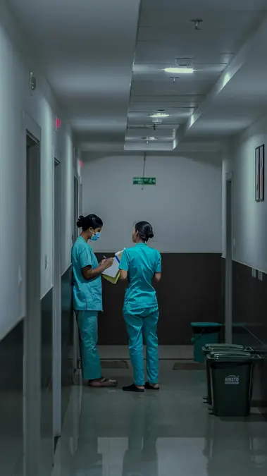 Two nurses in teal scrubs talking with a clipboard in a hospital corridor