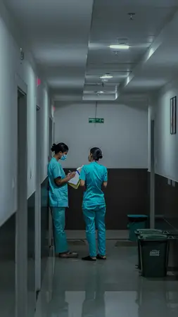 Two nurses in teal scrubs talking with a clipboard in a hospital corridor