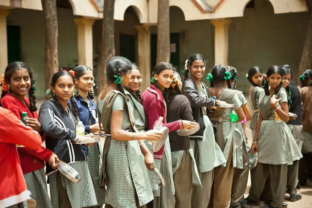 Schoolgirls in green checked uniforms standing in a line holding steel plates for a meal