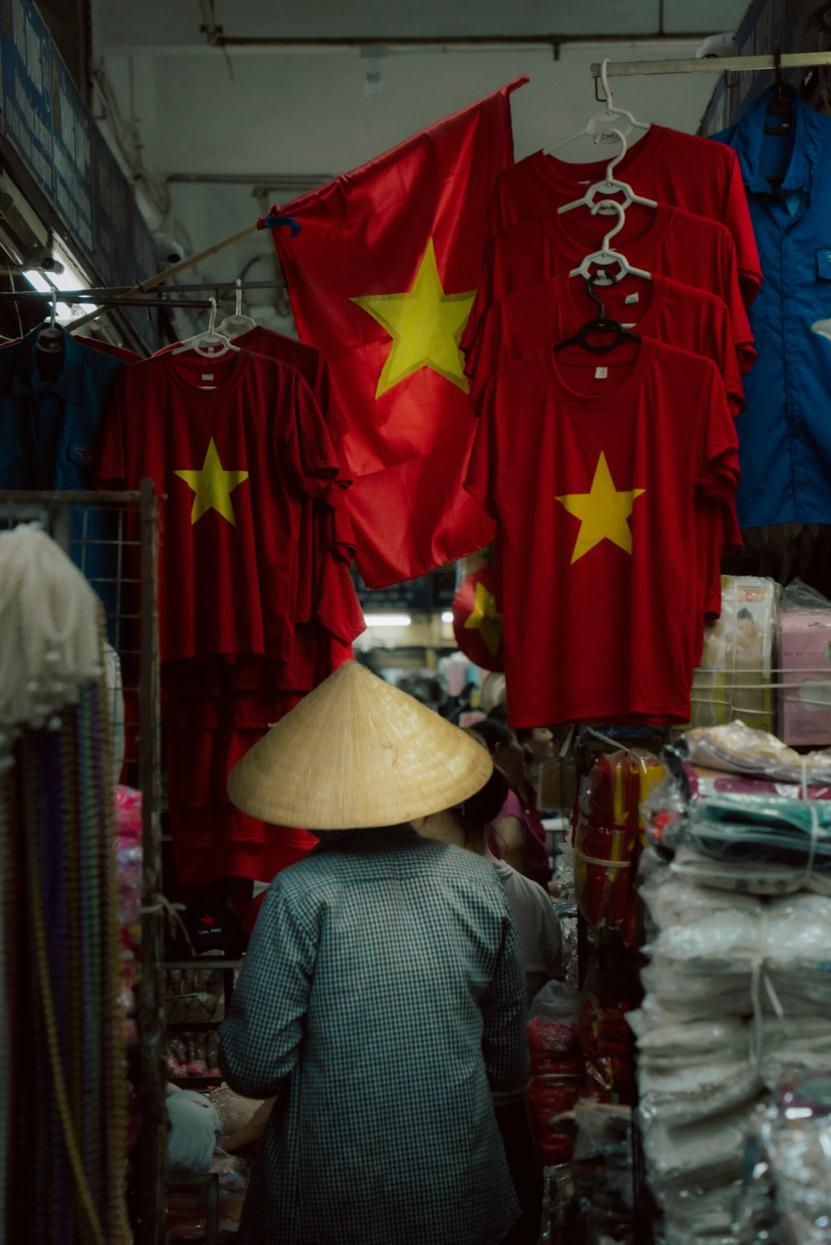 Vendor in a conical hat walking past red Vietnamese flag T-shirts hanging in a market stall