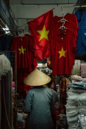 Vendor in a conical hat walking past red Vietnamese flag T-shirts hanging in a market stall