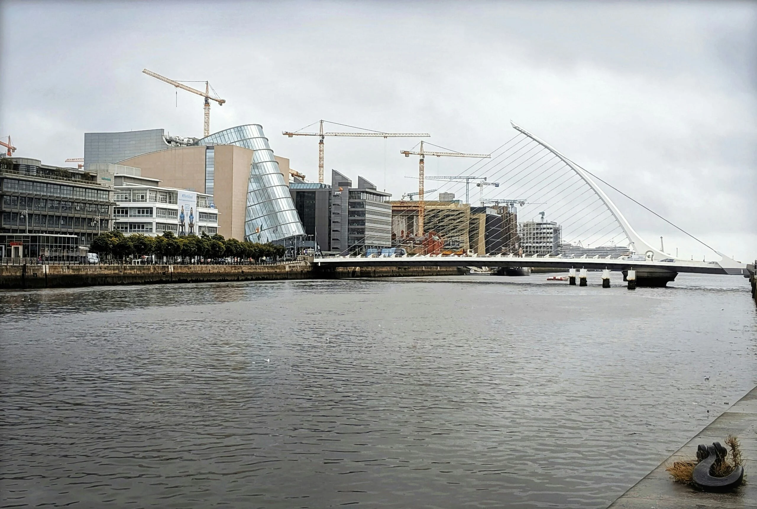 Dublin riverfront with cranes over glass office buildings and a white harp-shaped bridge under a grey sky