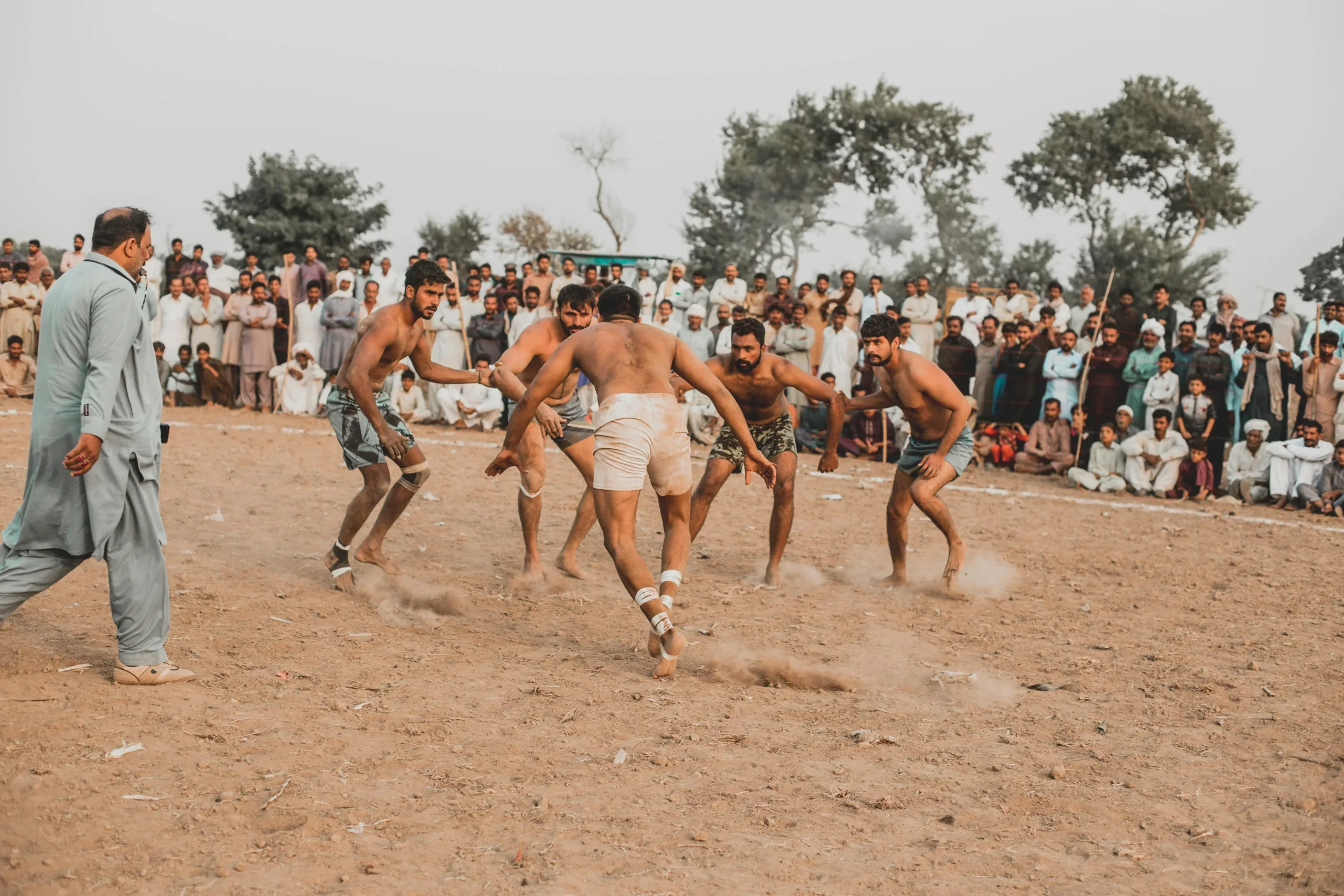 Shirtless kabaddi players on a dusty village field with a crowd of spectators seated around the edge and a referee at left