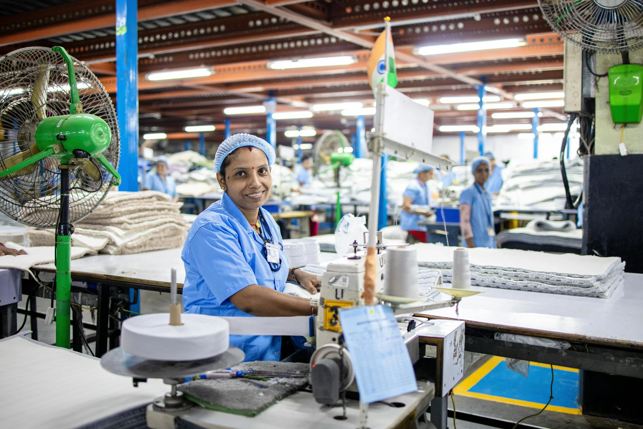 Woman in a blue uniform and hair cap smiling at a sewing station in a textile factory with fans and fabric stacks