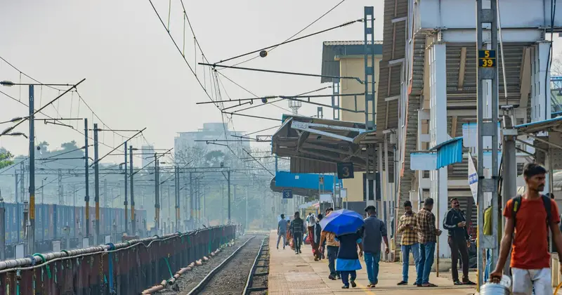 Railway platform with passengers walking, one under a blue umbrella, beside overhead wires and a track in morning haze