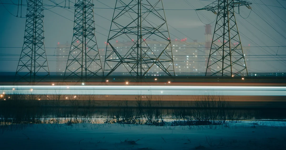 Electricity pylons and power lines at dusk with a blurred light trail and city lights behind