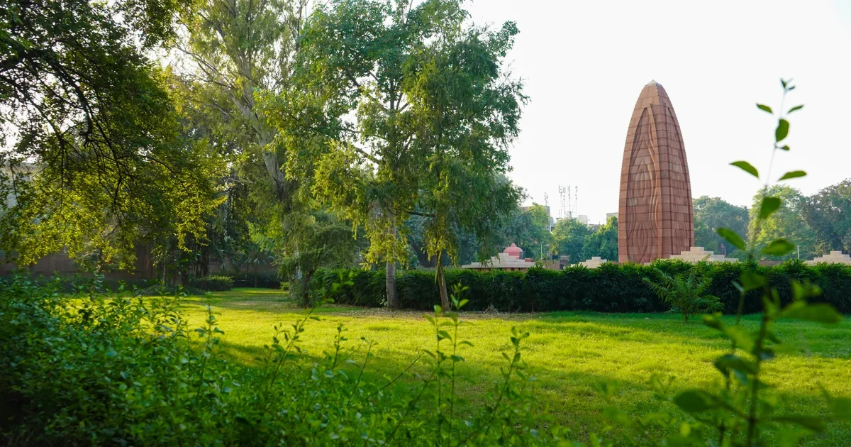 Tall red memorial tower rising behind trees and a green lawn in a garden