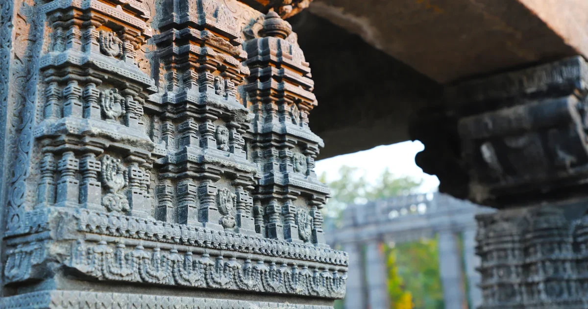 Close-up of a grey stone temple pillar carved in tiers of miniature shrines, with a colonnade blurred behind