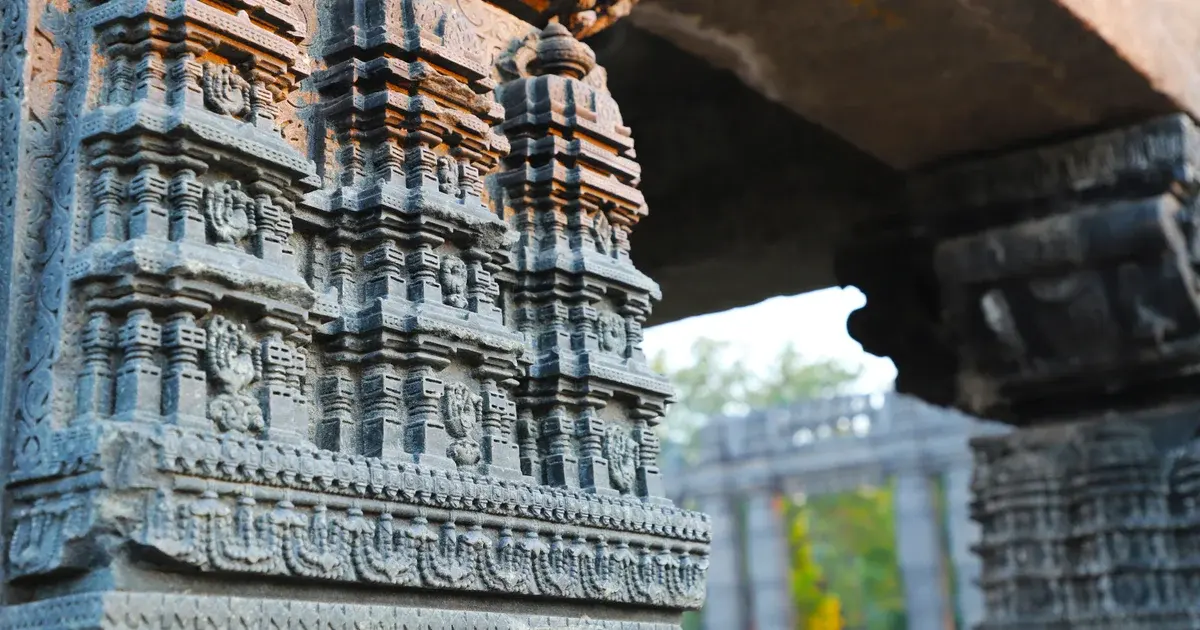 Close-up of a grey stone temple pillar carved in tiers of miniature shrines, with a colonnade blurred behind