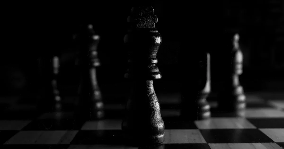 Black chess king in sharp focus on a chessboard with other dark pieces blurred behind it in low light