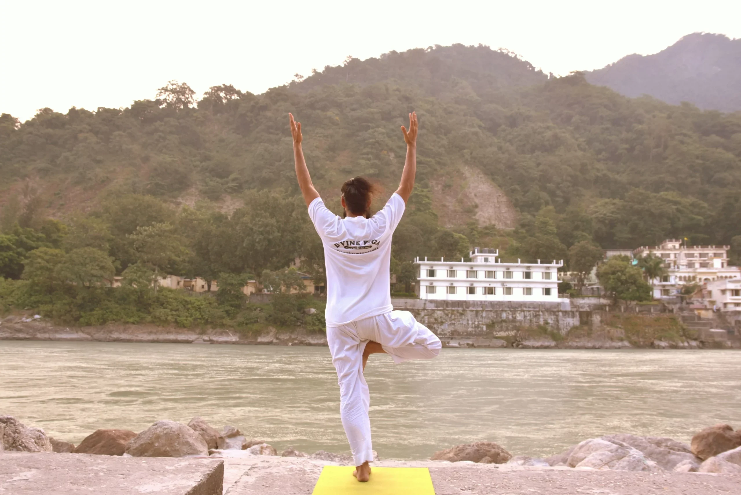 Man in white balancing in a tree pose on a yoga mat beside a river with forested hills behind
