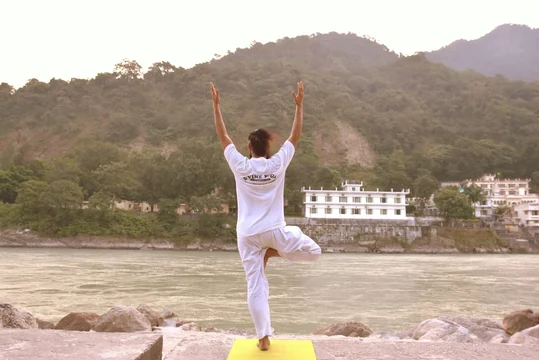 Man in white balancing in a tree pose on a yoga mat beside a river with forested hills behind