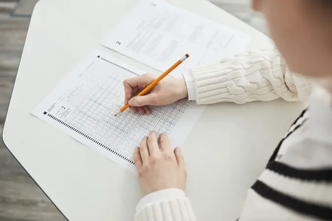Person's hands filling in an optical answer sheet with a pencil next to a question paper