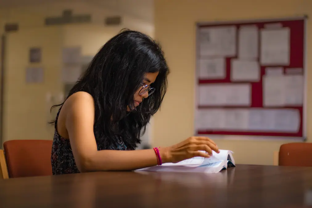 Young woman with glasses reading an open book at a table in a study room