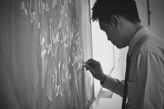 Black and white photo of a man in a shirt and tie writing equations on a chalkboard