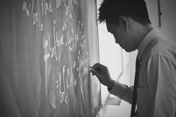 Black and white photo of a man in a shirt and tie writing equations on a chalkboard