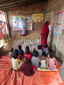 Two women teaching young children seated on mats in a small brick anganwadi room with wall posters