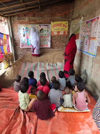 Two women teaching young children seated on mats in a small brick anganwadi room with wall posters