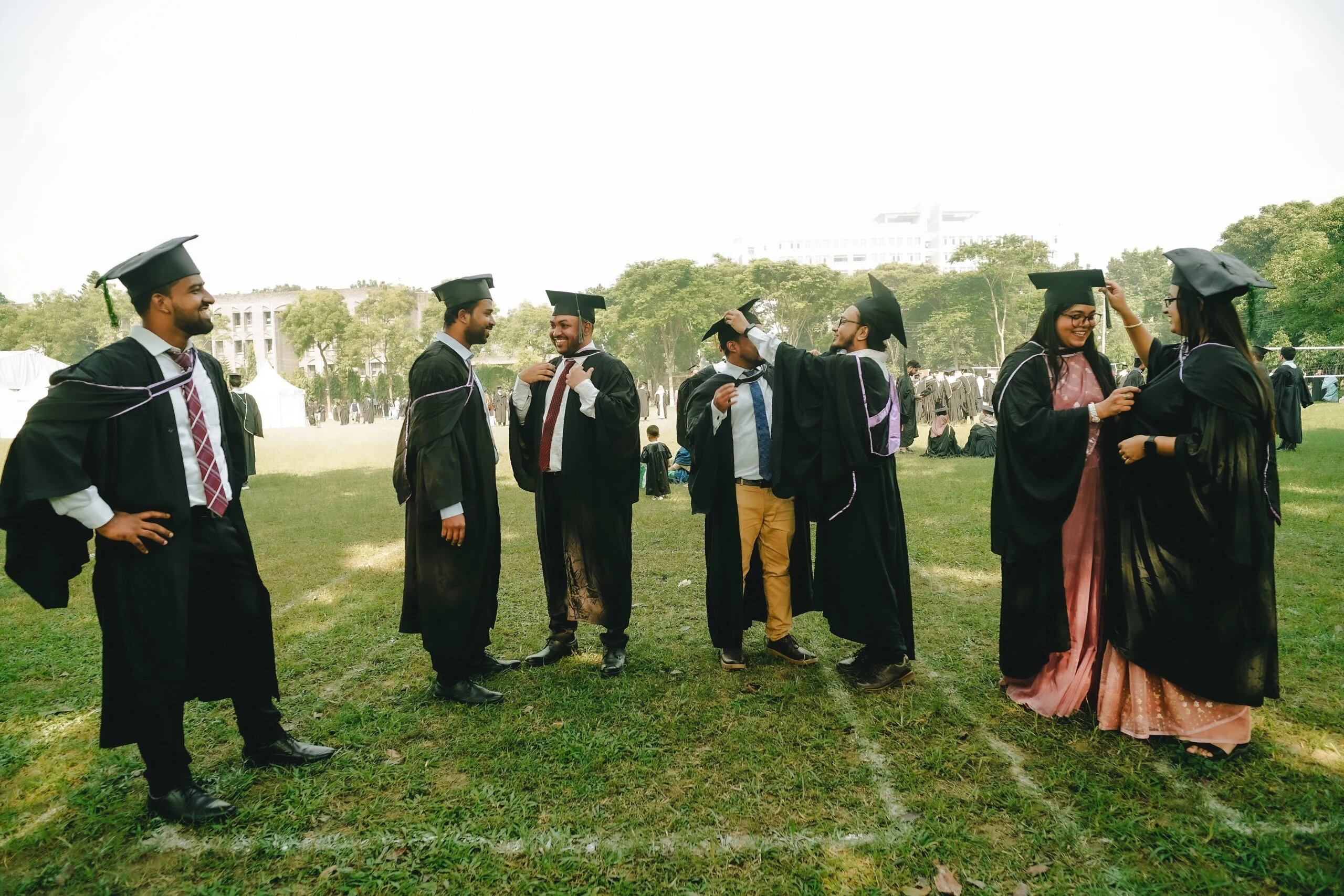 Graduates in black gowns and caps adjusting each other's robes on a grass lawn on campus