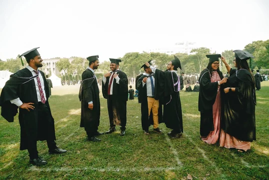 Graduates in black gowns and caps adjusting each other's robes on a grass lawn on campus