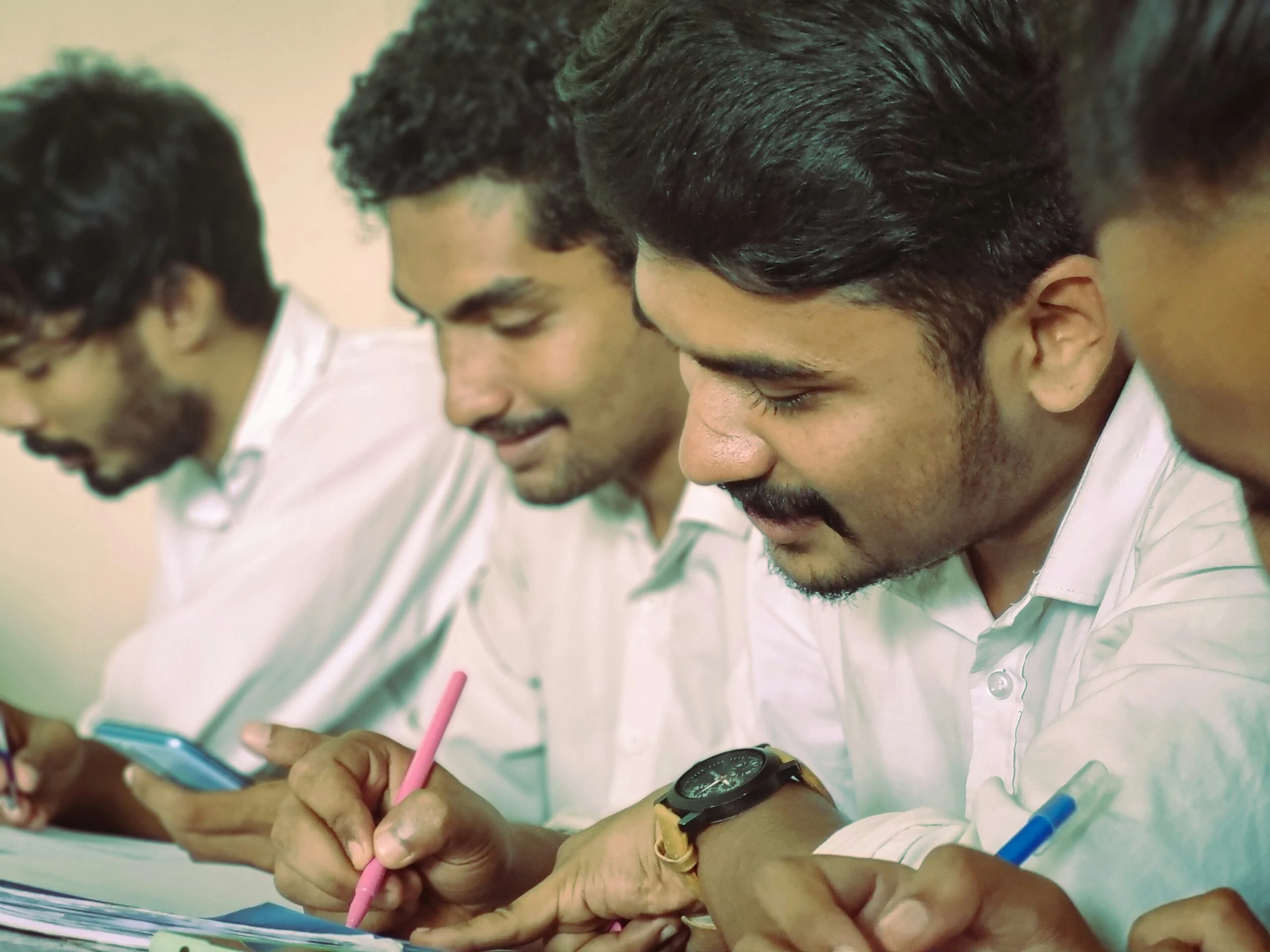 Young men in white shirts bent over desks writing with pens, one holding a phone