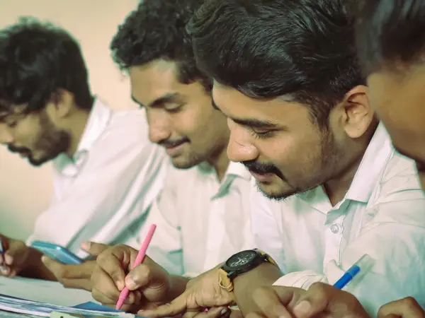 Young men in white shirts bent over desks writing with pens, one holding a phone