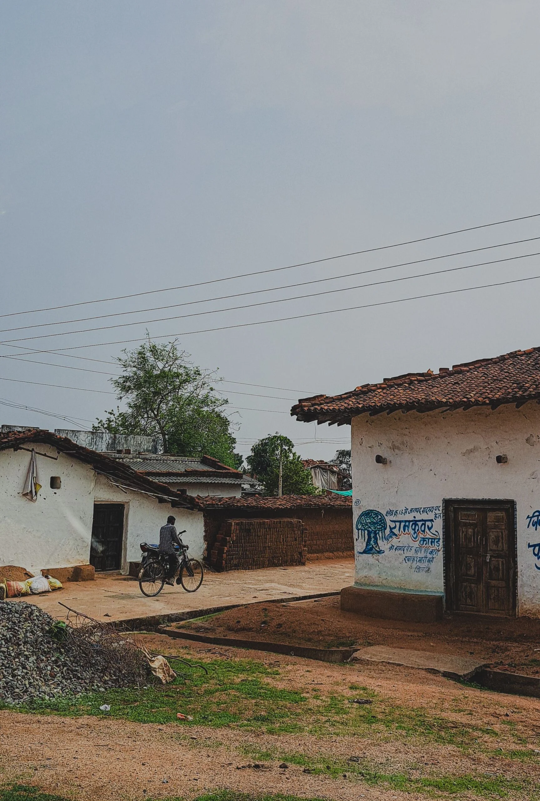 Village lane with whitewashed tiled-roof houses, a man on a bicycle and power lines under a grey sky