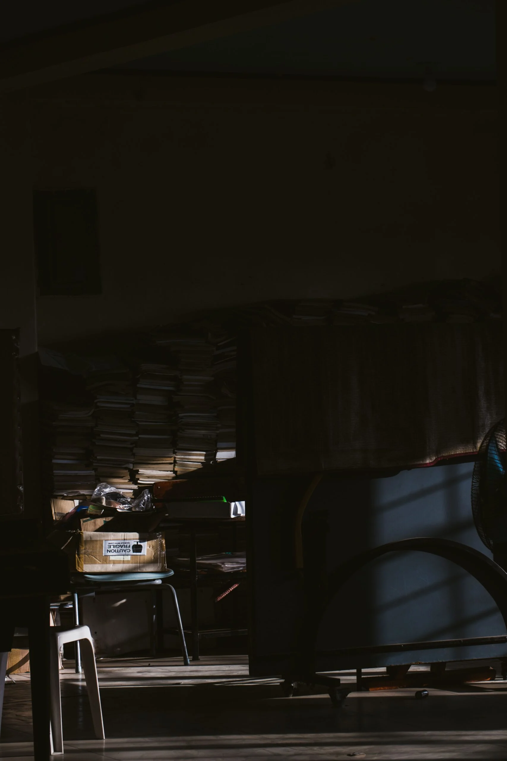 Dim room lit by a shaft of sunlight, with stacks of files and a cardboard box on a stool