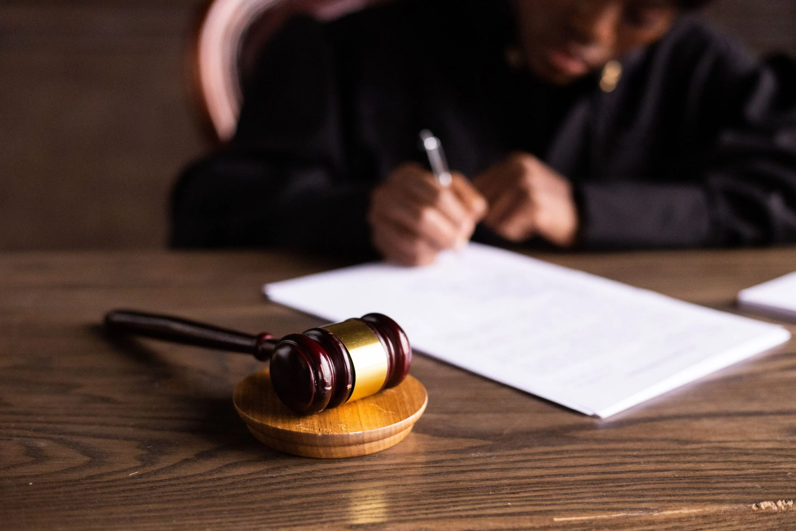 Wooden gavel on a desk with a blurred judge in a black robe signing papers in the background