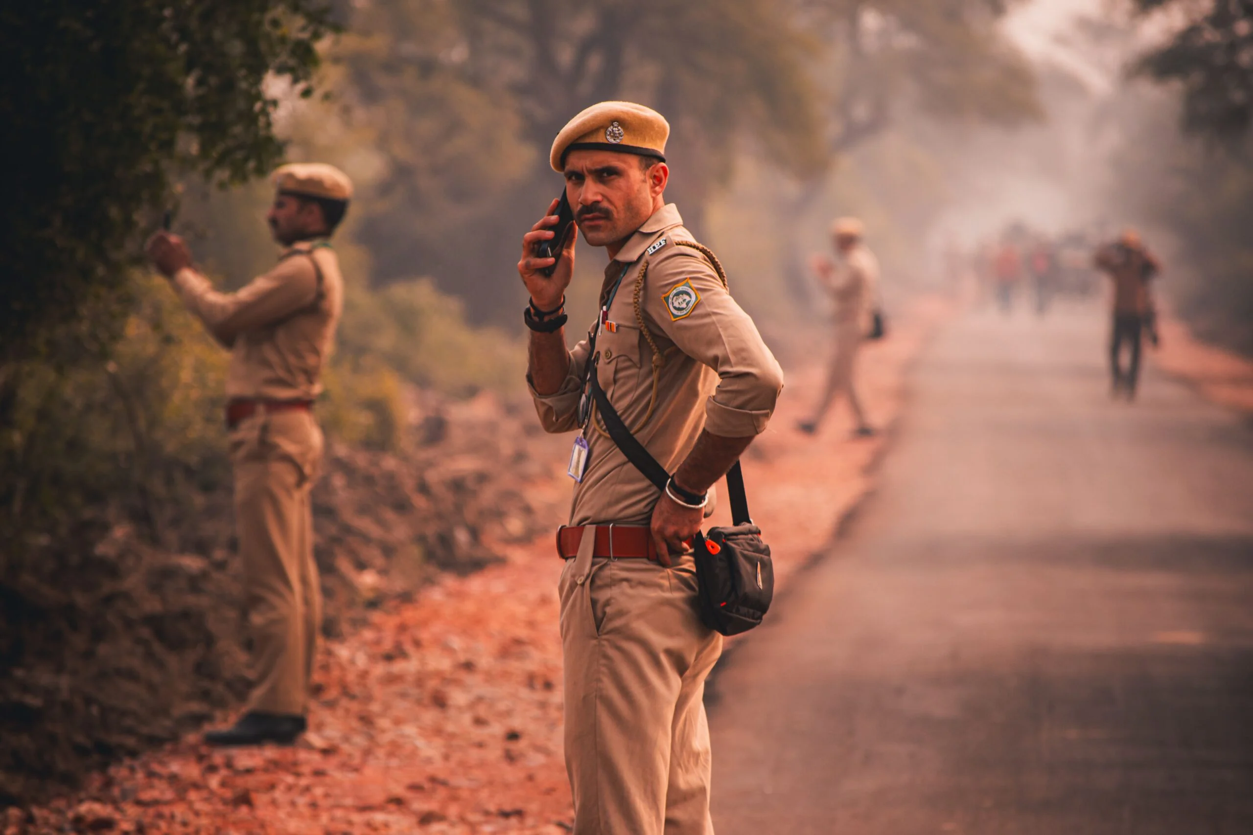 Uniformed police officers on a dusty road, one in the foreground speaking on a mobile phone