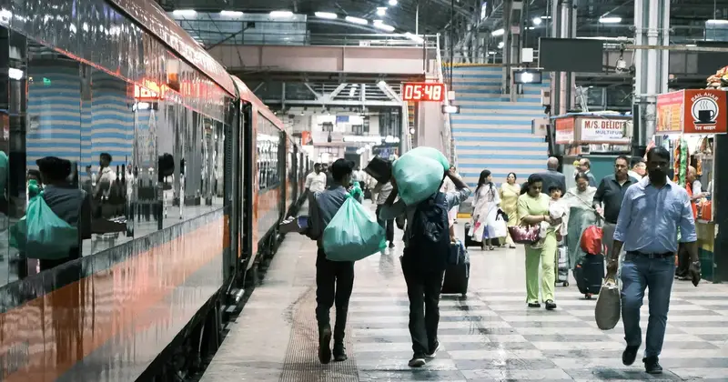 Passengers walking along a railway station platform beside a train, some carrying large green bundles on their heads
