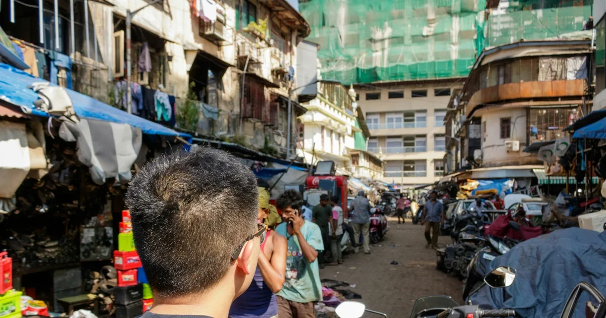 Crowded Indian street market with shop awnings, parked scooters, pedestrians and buildings behind