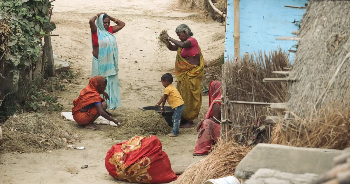 Women and a small child threshing straw and grain on a dusty village lane beside thatched huts