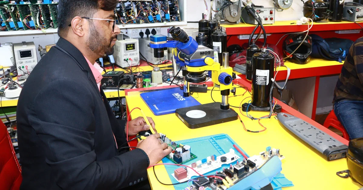 Repair technician in a dark blazer working on a circuit board at a yellow workbench with a microscope and tools