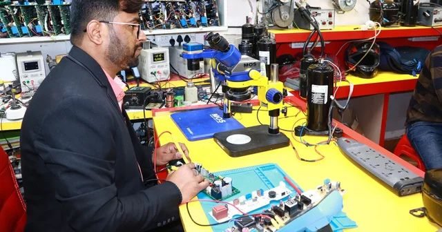 Repair technician in a dark blazer working on a circuit board at a yellow workbench with a microscope and tools