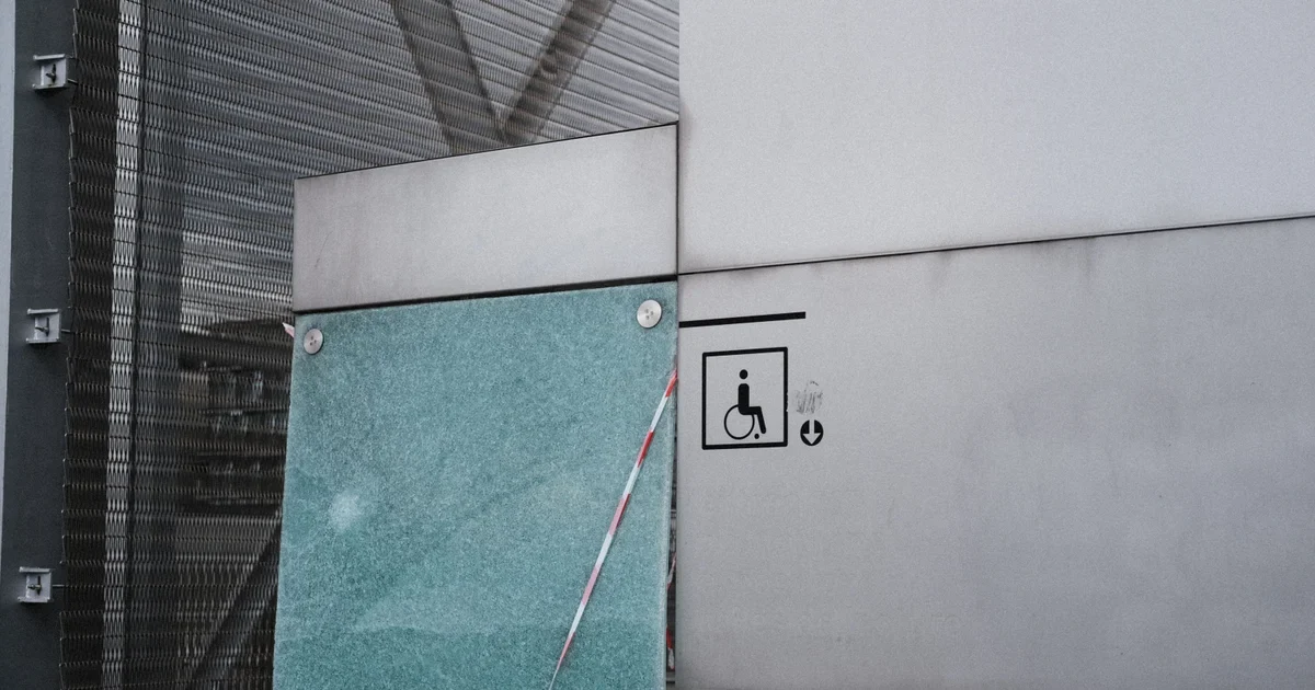 Wheelchair accessibility sign on a grey wall beside a cracked glass panel with red and white tape