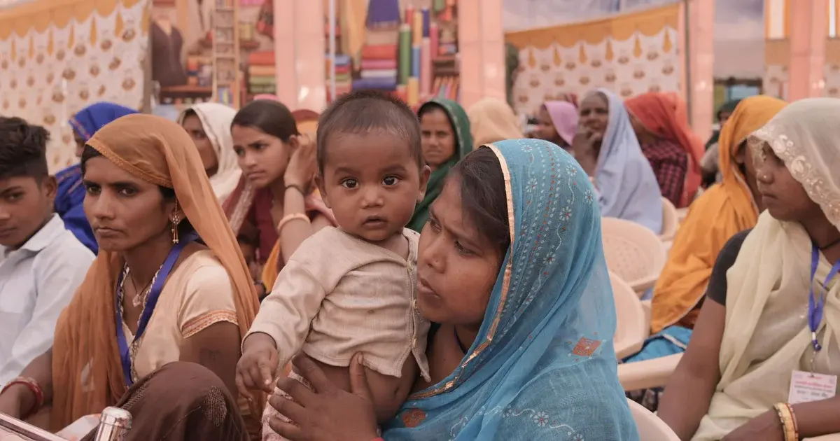 Woman in a blue sari holding a baby while seated among women at a community gathering