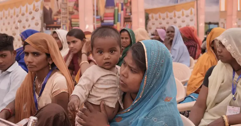 Woman in a blue sari holding a baby while seated among women at a community gathering