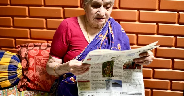 Elderly woman in a purple sari reading a newspaper in front of an orange brick wall