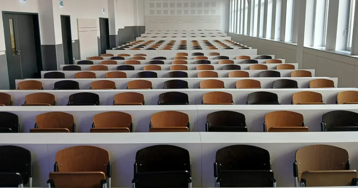 Rows of empty wooden lecture hall seats with long white desks stretching to the back of a large room