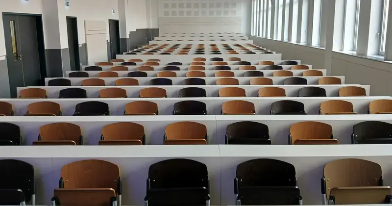 Rows of empty wooden lecture hall seats with long white desks stretching to the back of a large room