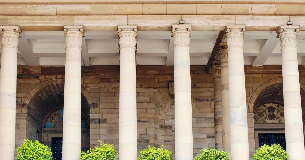 Sandstone building facade with tall columns and arched doorways behind small green shrubs