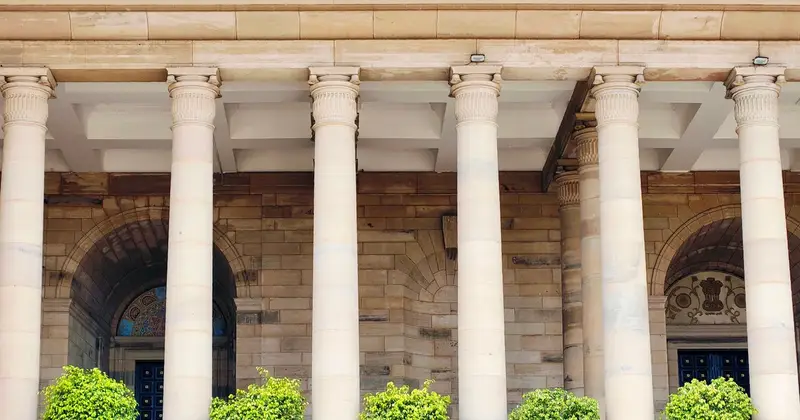 Sandstone building facade with tall columns and arched doorways behind small green shrubs