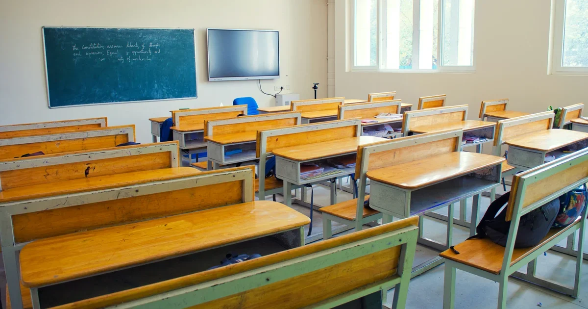 Empty classroom with wooden desks and benches facing a green chalkboard and a wall-mounted screen