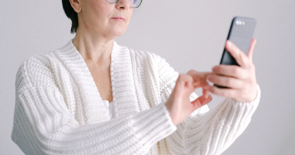 An older woman in glasses and a white sweater looking at her smartphone at home