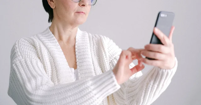 An older woman in glasses and a white sweater looking at her smartphone at home